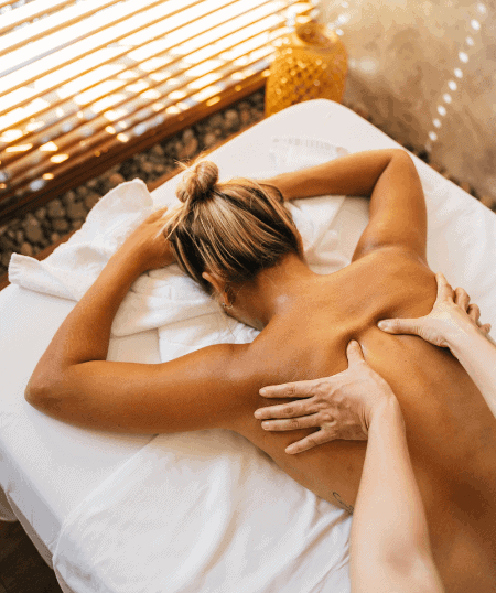 A person is lying face down on a massage table with a white towel. Another person's hands are skillfully administering a shiatsu massage to their back. There's soft lighting and a window with blinds in the background.