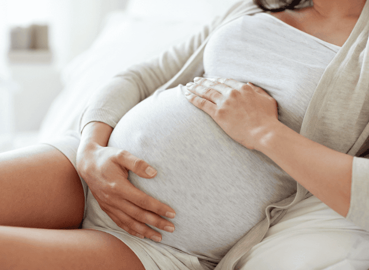 A pregnant woman in a light gray outfit sits on a couch with her hands gently resting on her baby bump, perhaps contemplating the soothing relief of a pregnancy massage.