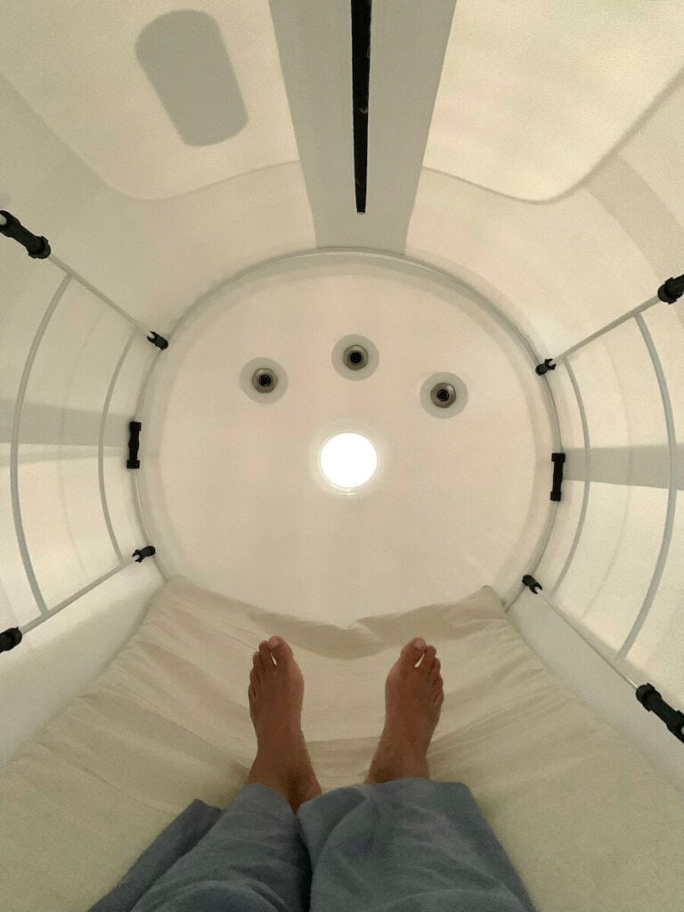 A person lying inside a white, enclosed hyperbaric chamber, seen from the feet end, with three circular vents and a circular light on the ceiling.