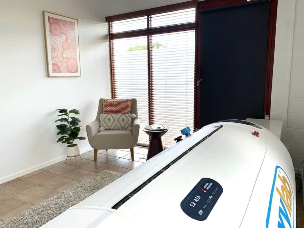 A brightly lit room with a massage chair, a potted plant, framed artwork, and a massage bed near a large window with blinds.