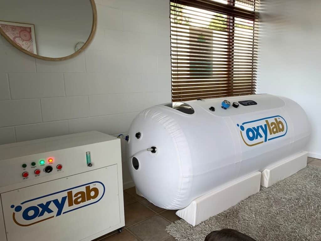 A white hyperbaric oxygen therapy chamber labeled "oxylab" is positioned in a room with large windows and wooden blinds. A control unit connected to the chamber is on the left.