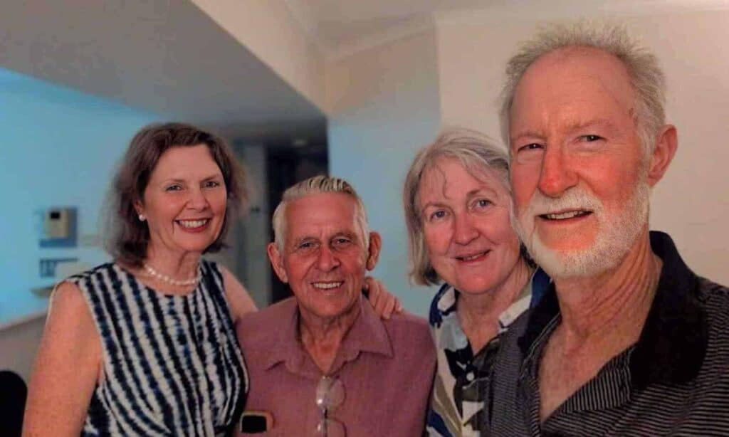 Four older adults, two men and two women, stand close together indoors, smiling at the camera.