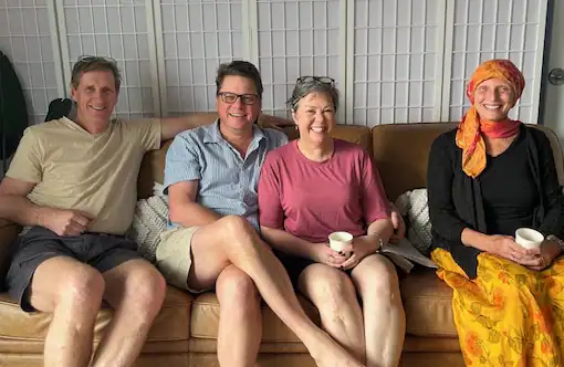 Four adults sit together on a brown couch, smiling at the camera. Three hold mugs, and one wears a colorful headscarf and skirt. A white paneled screen is in the background.